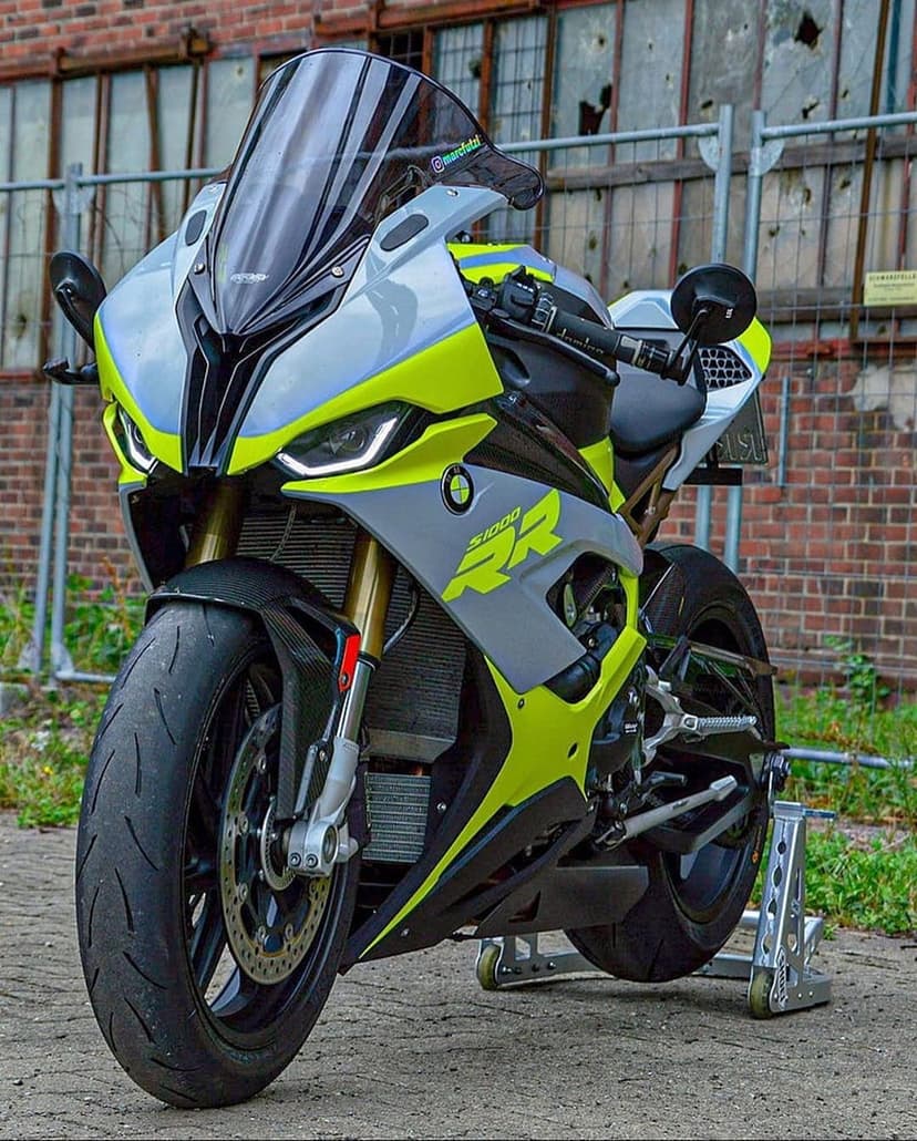 BMW S1000RR superbike with grey and lime green fairings stands against a weathered brick wall on a paddock stand.