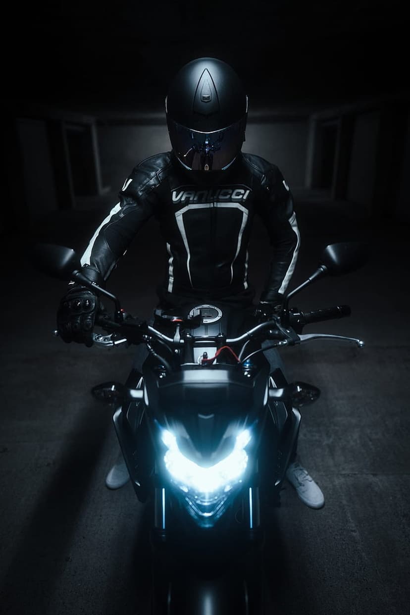 Motorcyclist in black leather gear sits on a bike with two bright circular headlights shining in the dark.