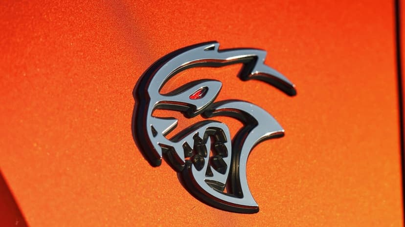 Chrome Hellcat emblem with a glowing red eye mounted on a vibrant metallic orange car surface with sharp highlights.