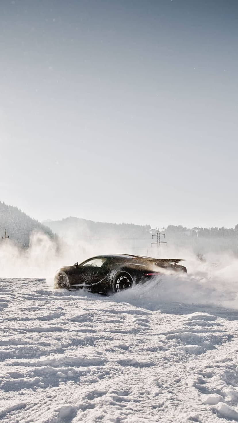 Black Bugatti supercar drifting on a snowy road with white powder spraying near pine trees and distant mountains.