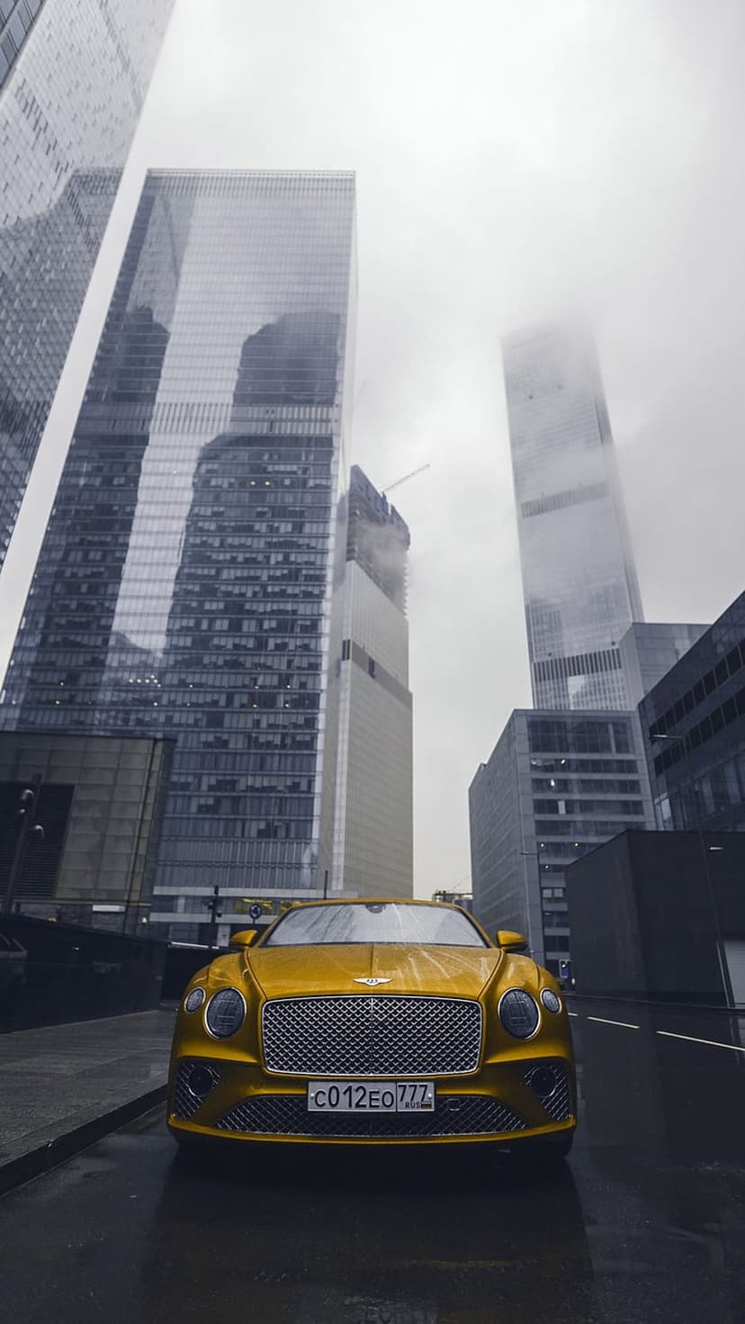 Golden Bentley Continental GT on a rain-slicked city road surrounded by modern glass skyscrapers and clouds.