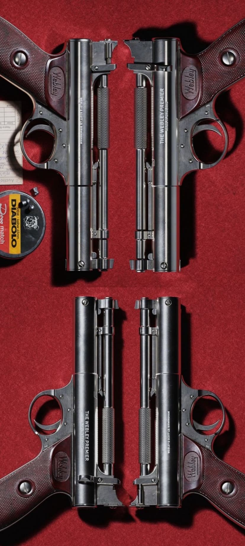 Two Webley Premier air pistols symmetrically arranged on red felt with a pellet tin and loose metal pellets.