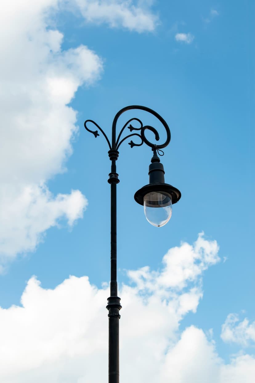 Ornate black wrought-iron street lamp with decorative scrolls silhouetted against a bright blue sky with clouds.