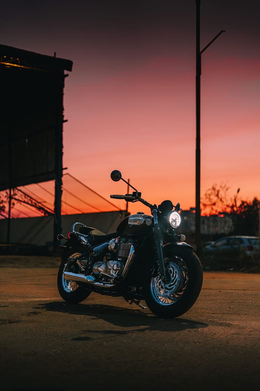 Black vintage motorcycle with a glowing headlight parked on asphalt under a vibrant orange and pink sunset sky.