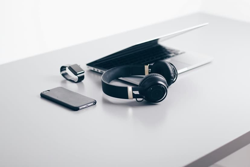 Black laptop, over-ear headphones, smartphone, and smartwatch resting on a clean grey desk in a flat lay view.