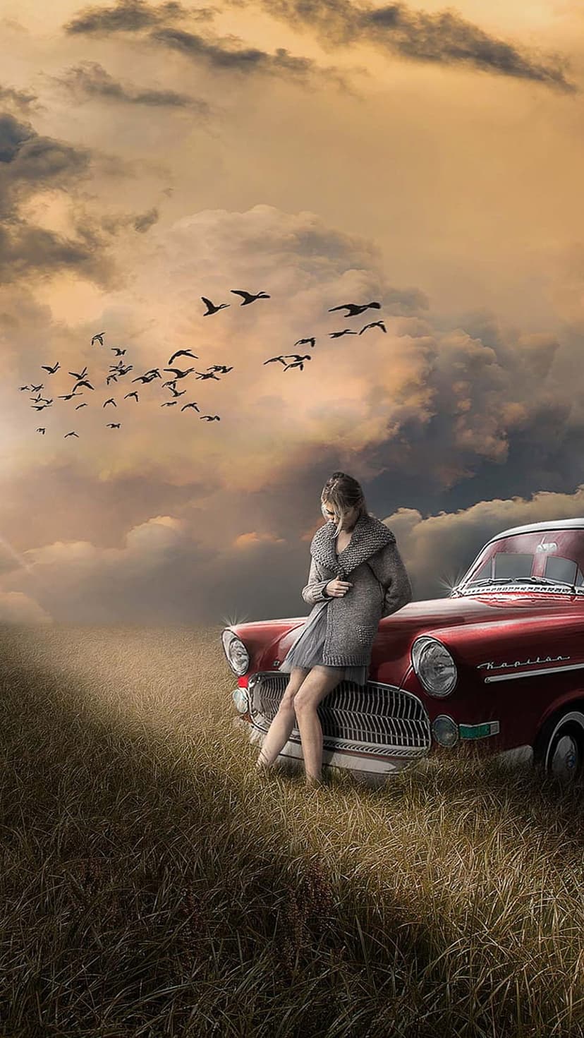 Woman in a grey cardigan leaning on a red vintage car while birds fly through a sunset sky with clouds.