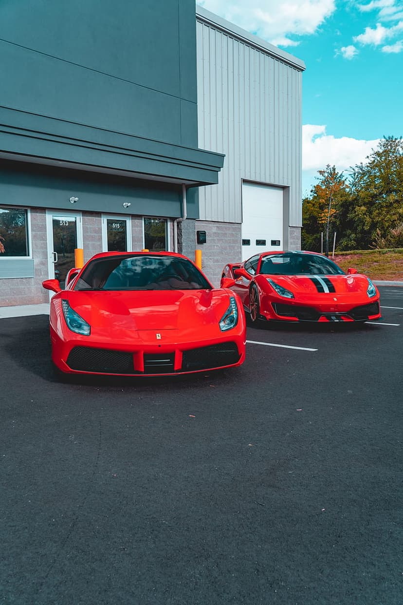 Two red Ferrari sports cars parked in front of a modern grey commercial building under a bright blue sky.