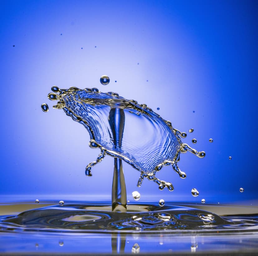 Two water droplets collide into a blue umbrella shape with tiny beads of liquid scattering in mid-air.