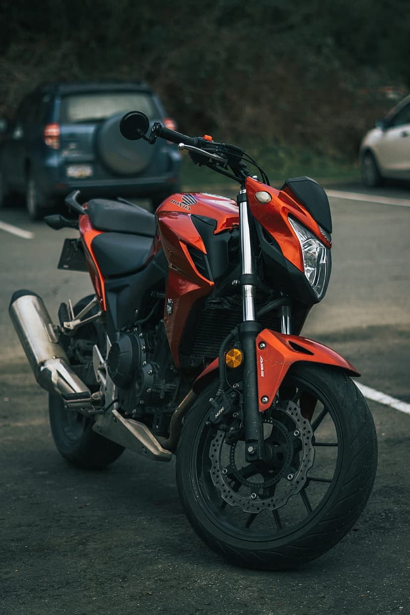 Orange Honda motorcycle parked on an asphalt road with visible disc brakes, exhaust pipe, and background cars.