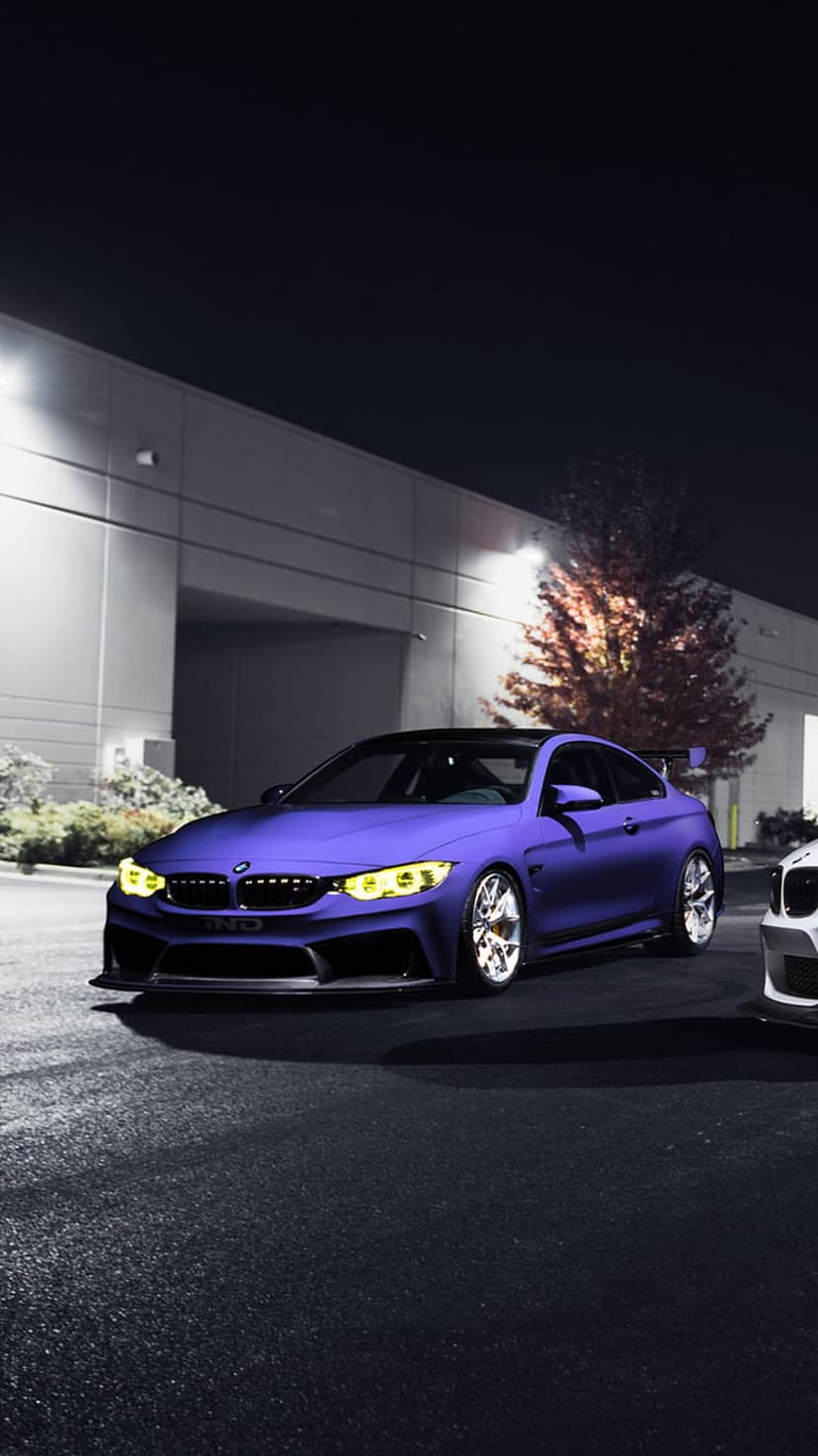 Purple BMW M4 Coupe with yellow headlights parked on a wet street at night in front of a modern lighted building.