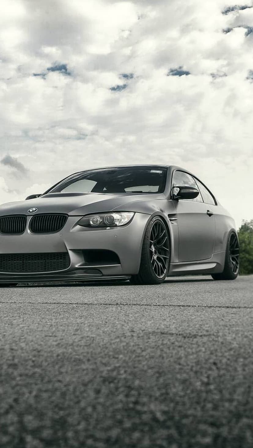 Matte gray BMW M3 coupe with black wheels and a lowered stance on asphalt against a dark overcast sky backdrop.
