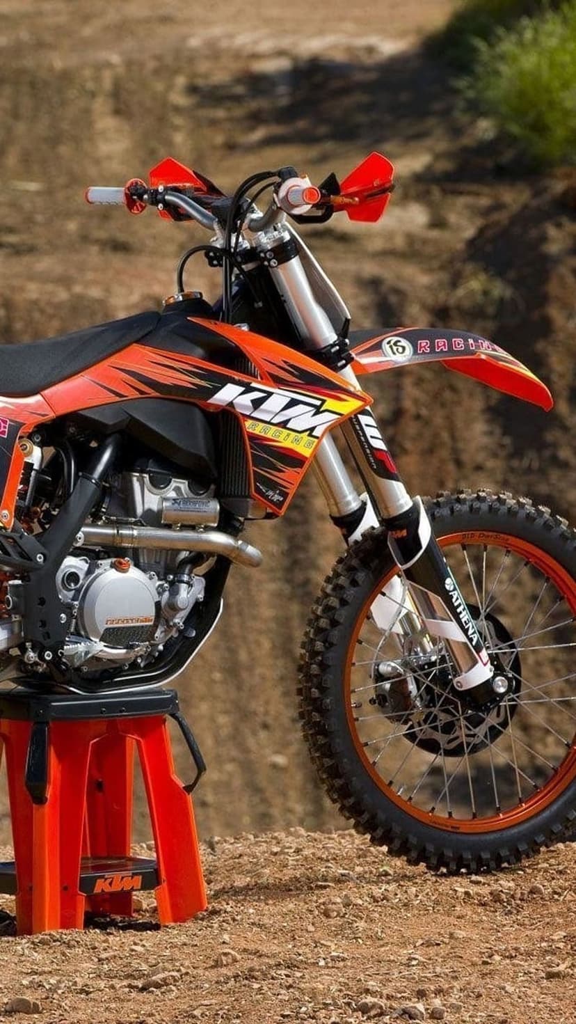 KTM motocross bike with orange frame and black decals sits on a matching stand against a dusty terrain backdrop.