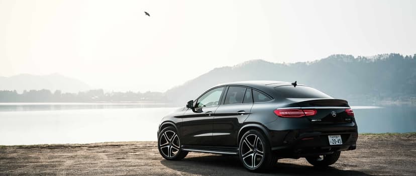 Black Mercedes-Benz GLE Coupe parked on a gravel shore by a calm lake with misty mountains in the background.