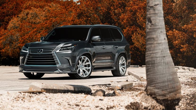 Charcoal Lexus LX SUV with custom silver rims parked in front of orange autumn trees on a paved lot.