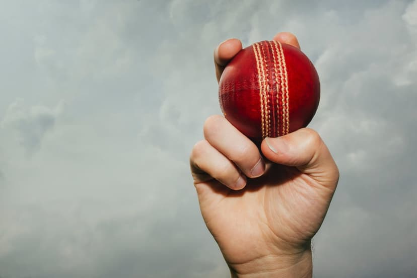 Hand gripping a red leather cricket ball with visible white stitching against a backdrop of grey storm clouds.