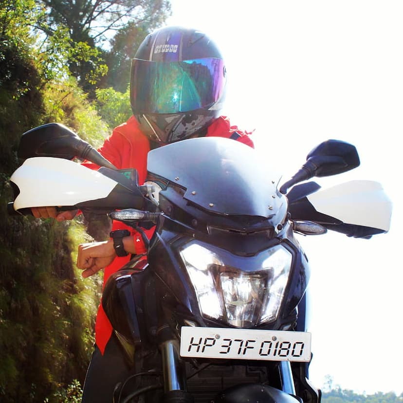Motorcyclist in a red jacket and silver helmet sits on a black bike against a blurred green background.