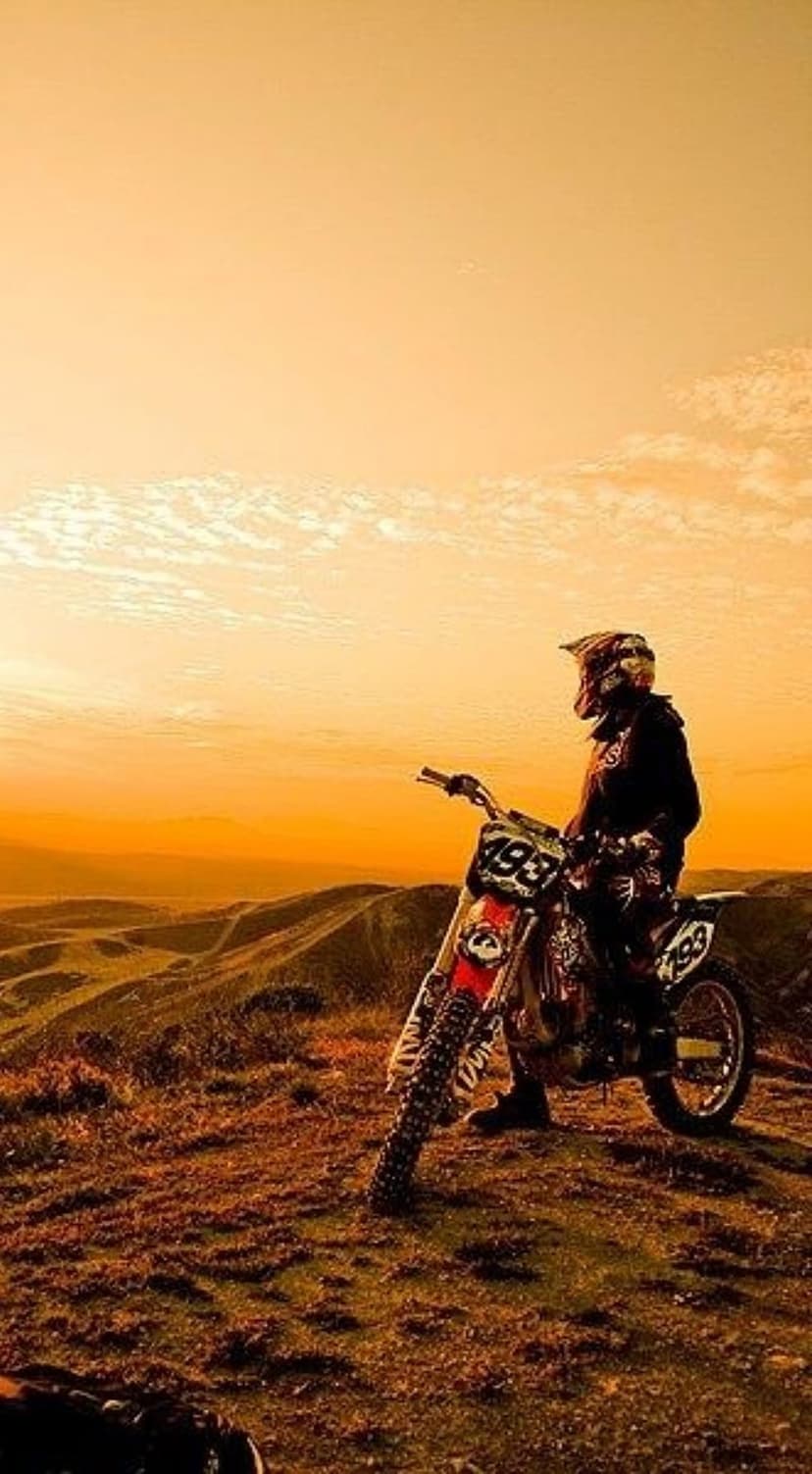 Motocross rider on a dirt bike silhouetted against an orange sunset sky atop a dusty desert mountain ridge.