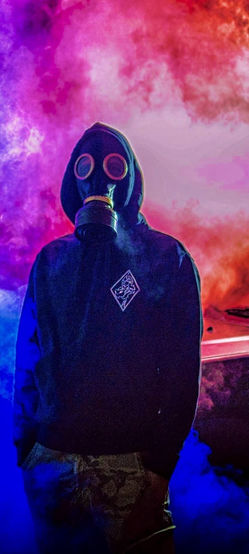 Person in a black hoodie and gas mask stands amidst dense clouds of pink, purple, and red neon smoke.