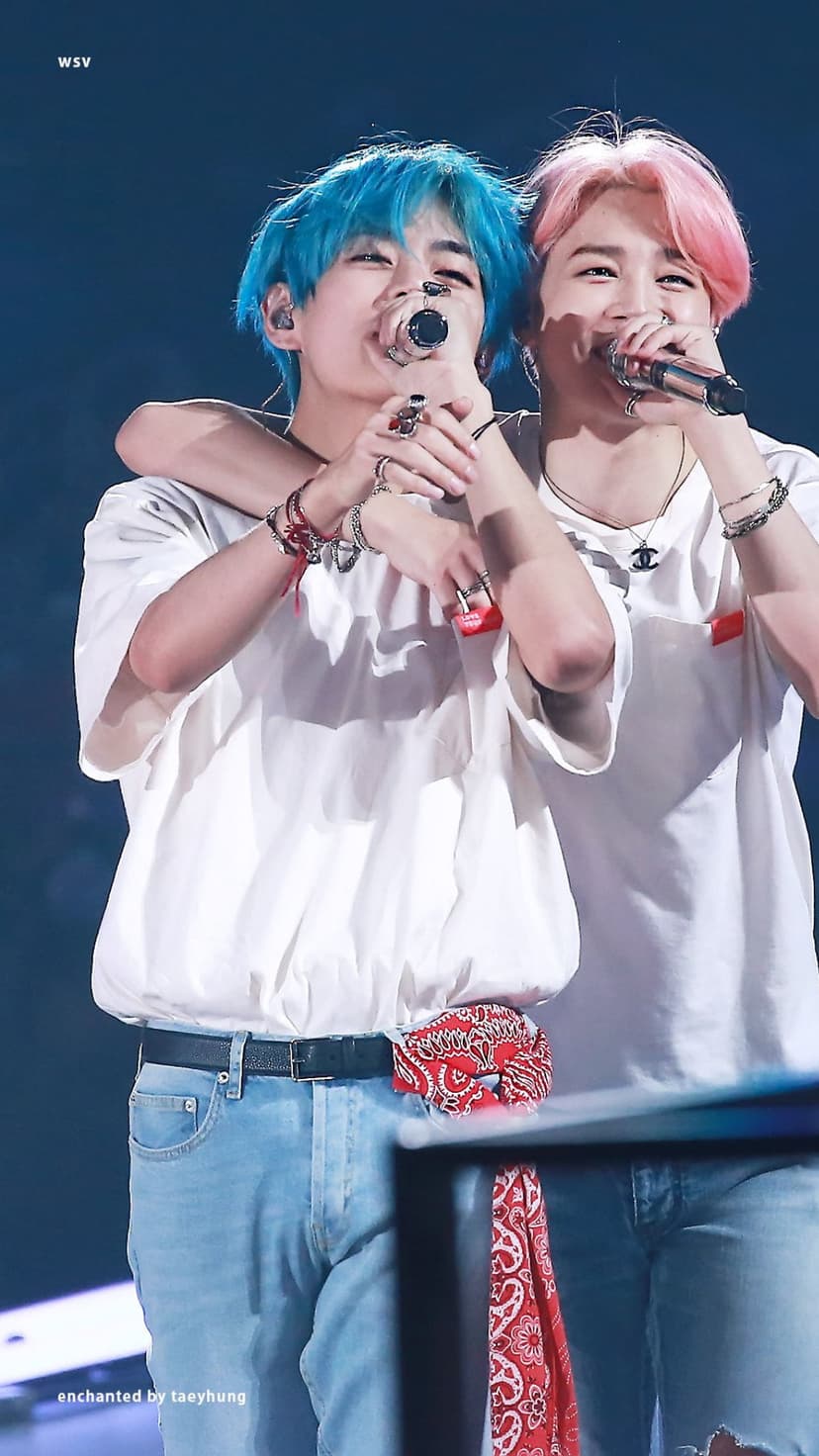 Taehyung with blue hair and Jimin with pink hair hug on stage while holding microphones and wearing white shirts.