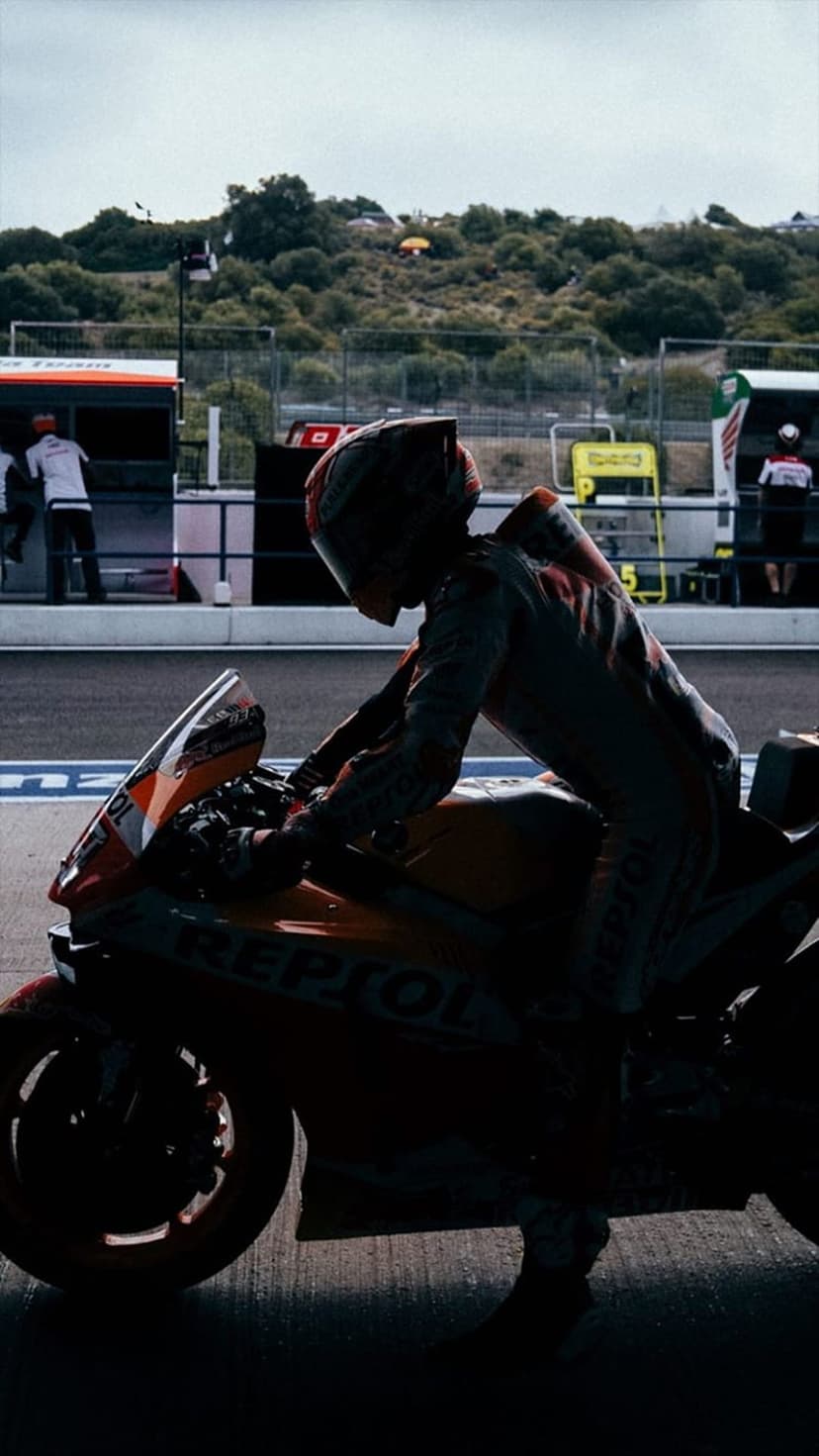 Motorcycle racer in silhouette sits on a Repsol bike in a sunny pit lane with grassy hills in the distance.