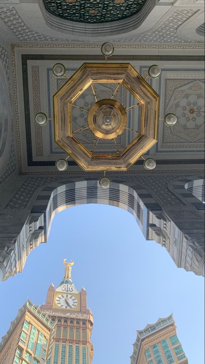 Golden chandelier and intricate mosque ceiling patterns frame the Makkah Clock Tower under a bright blue sky.