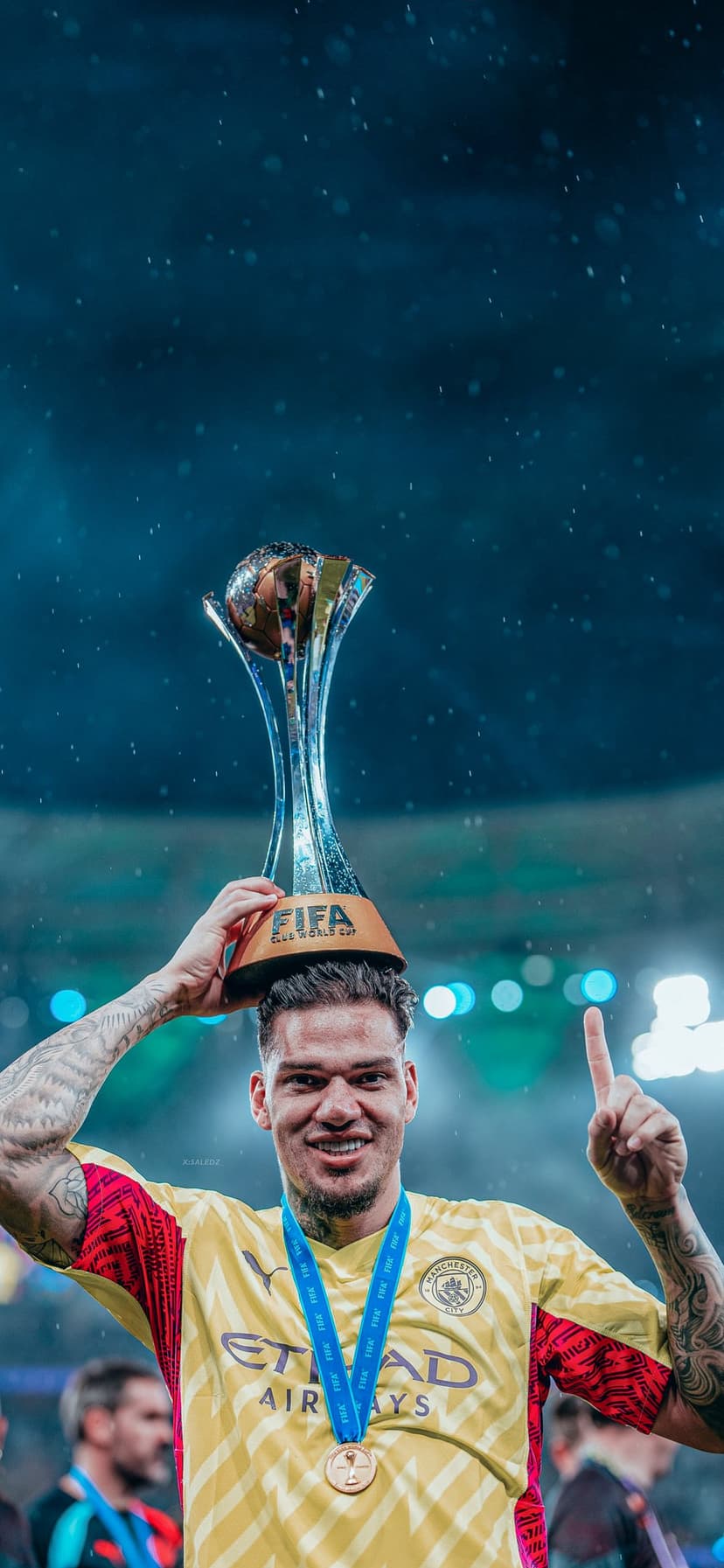 Manchester City goalie Ederson lifts a gold trophy wearing a yellow uniform and medal during a stadium celebration.