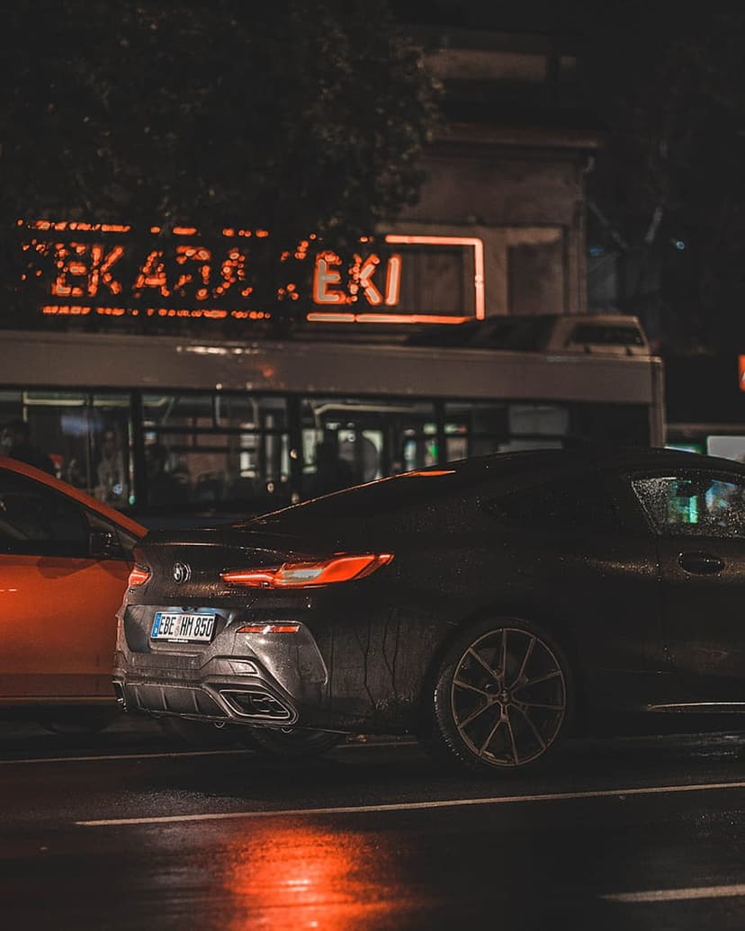 Rear view of a black BMW coupe on wet asphalt with glowing taillights and a neon Cyrillic sign in the background.