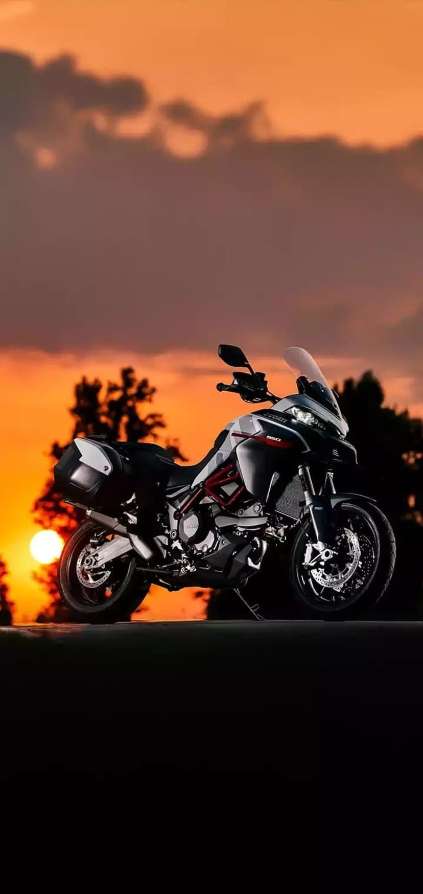 Ducati motorcycle silhouette stands before a glowing orange sunset with wispy clouds and dark tree shapes.