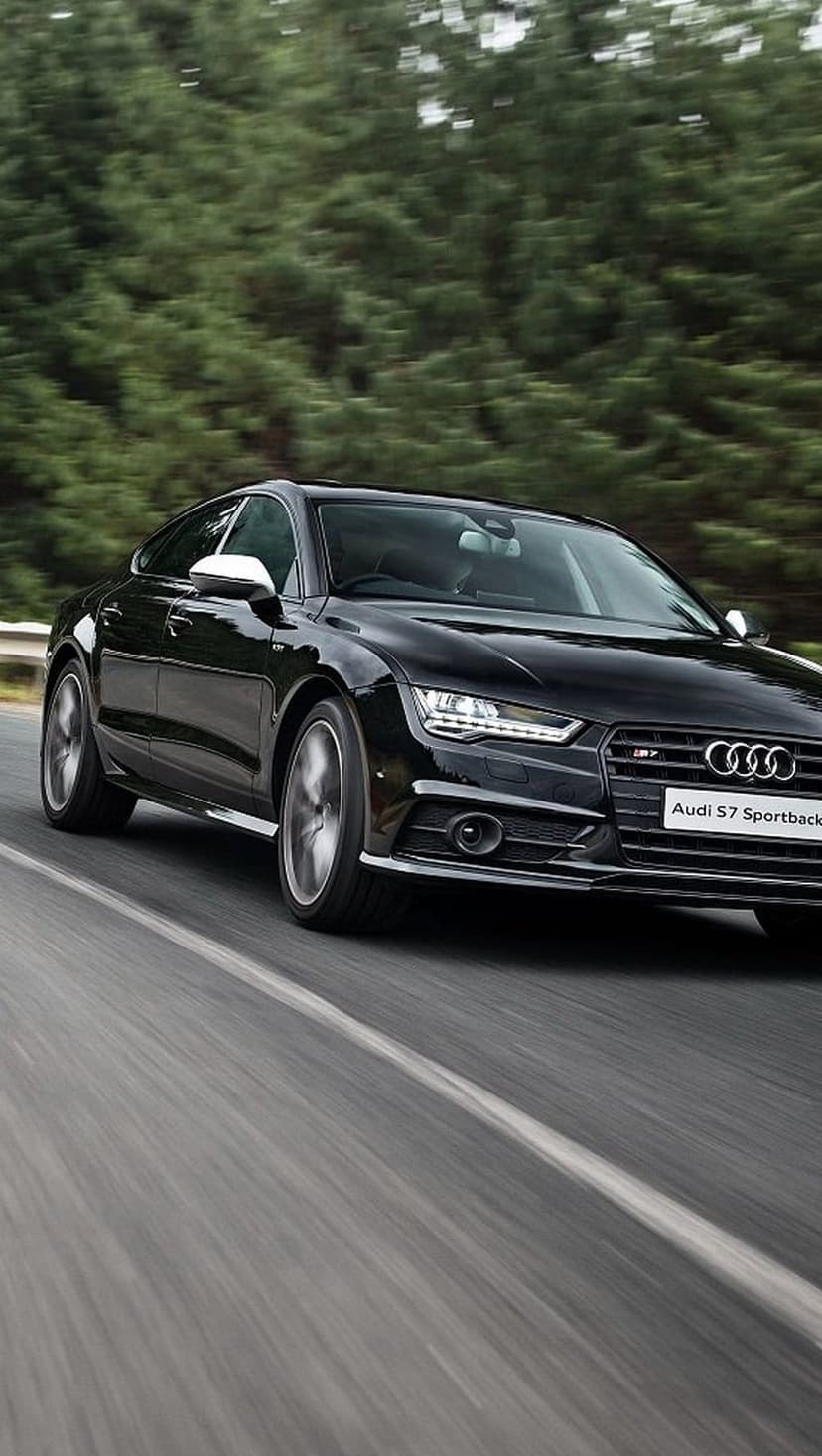 Black Audi S7 Sportback with glowing headlights driving on a paved road surrounded by blurred green forest trees.