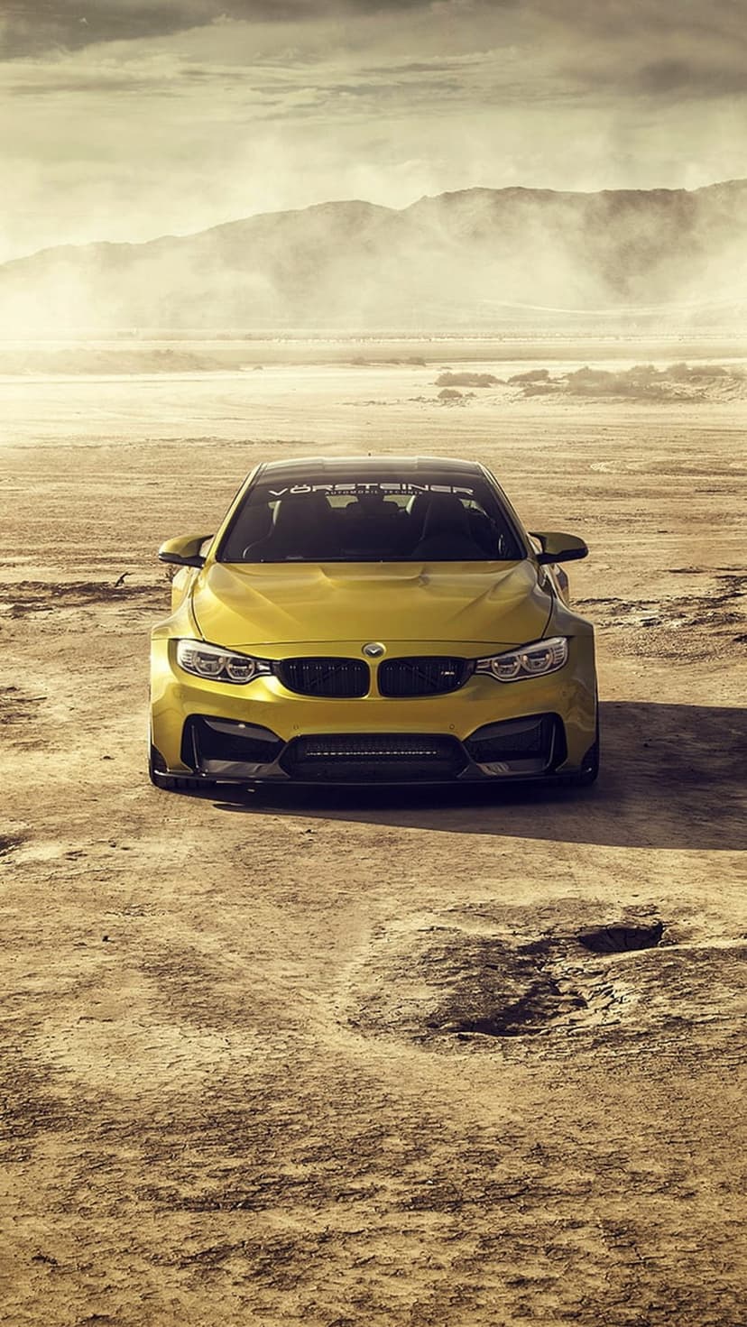 Yellow BMW M4 performance car with Vorsteiner styling parked on a dusty road with mountains under a cloudy sky.