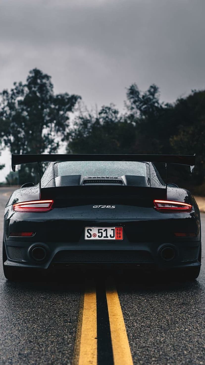 Black Porsche 911 GT2 RS rear view on wet asphalt with yellow lines under a cloudy sky in a vertical shot.