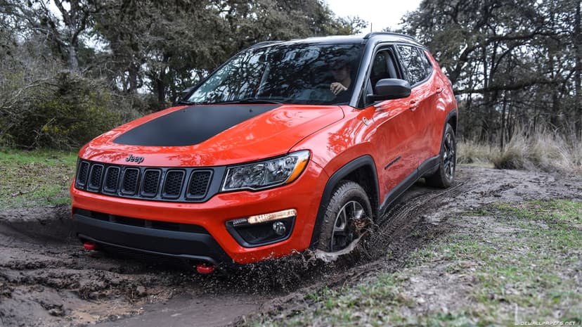Red Jeep Compass Trailhawk driving through a deep mud puddle on a wooded trail with mud splashing from the front tire.