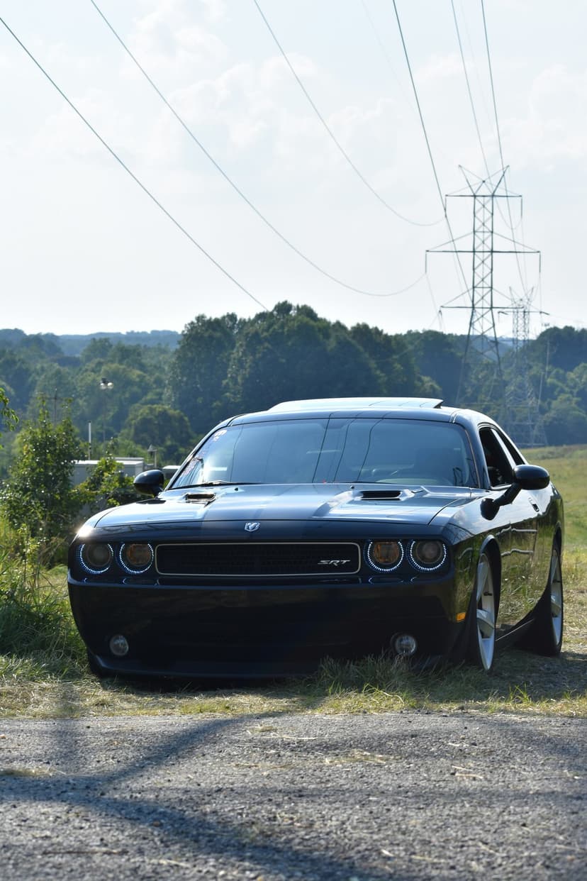 Black Dodge Challenger SRT parked on a gravel road with green trees and power lines under a clear sky.