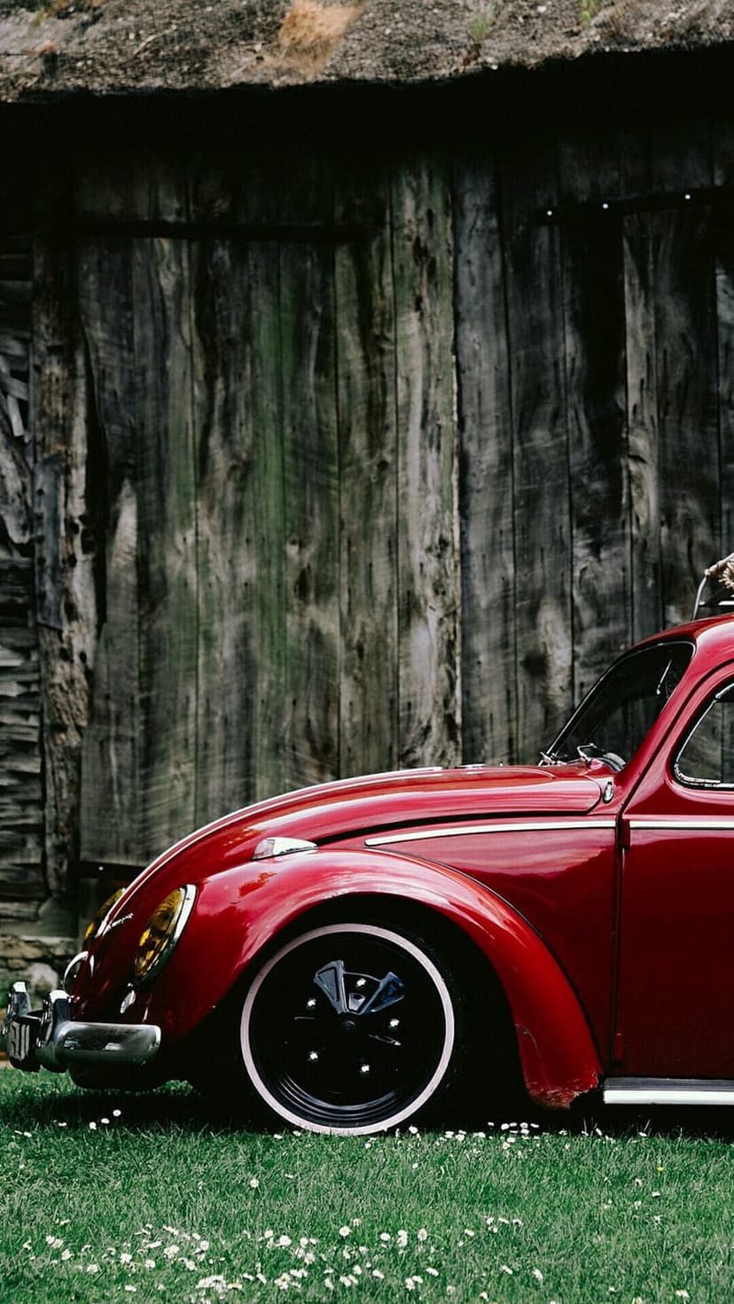 Red Volkswagen Beetle with yellow headlights parked on green grass in front of a weathered rustic wooden barn.
