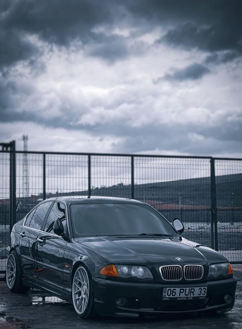 Dark BMW sedan with alloy wheels parked near a metal fence under a heavy, overcast sky during twilight hours.