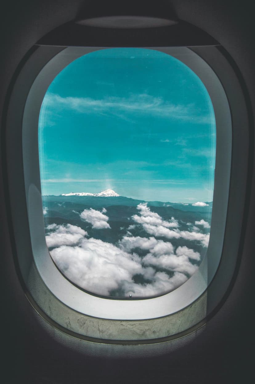 Snow-capped mountain peaks emerge from thick white clouds viewed through the curved frame of an airplane window.