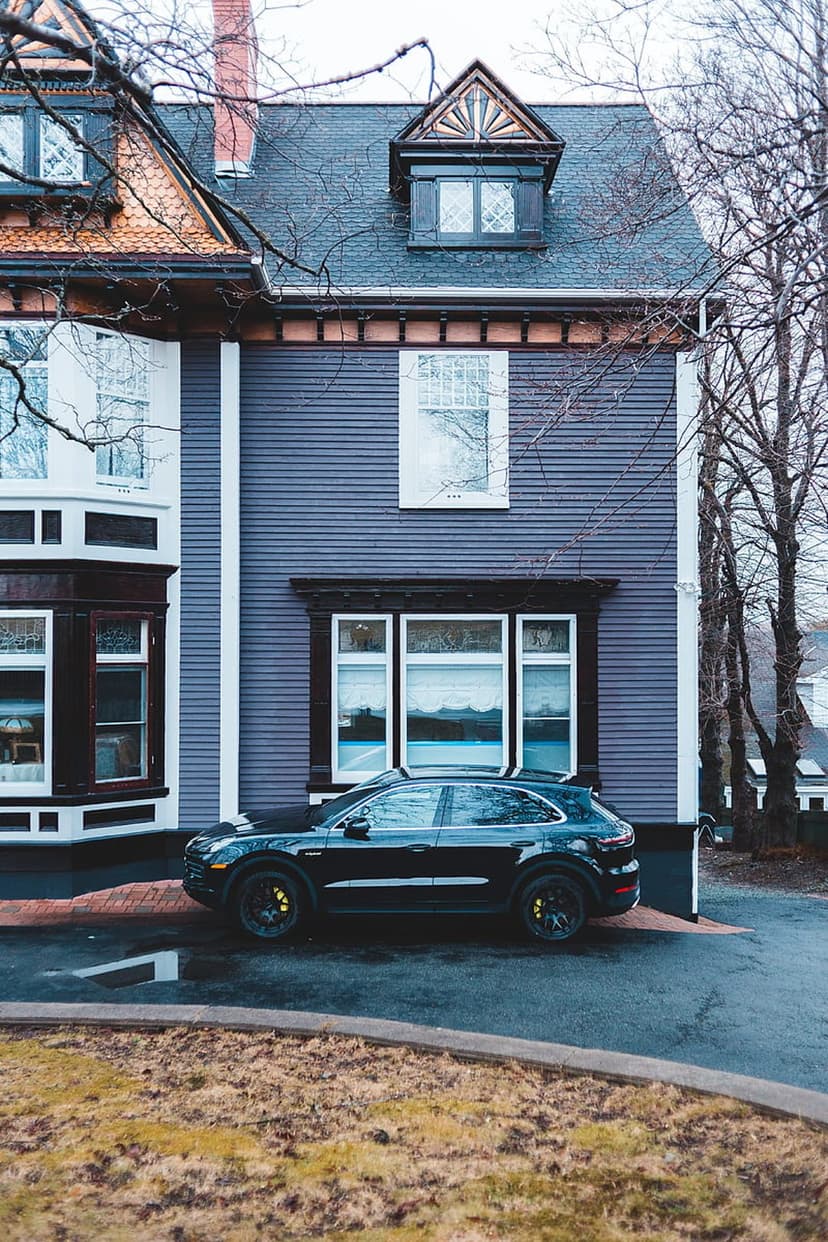 Black Porsche SUV parked on a wet asphalt driveway in front of a grand blue house with historic architecture.