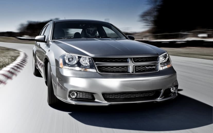Silver Dodge Avenger sedan speeding through a racetrack curve with a driver in a white helmet and motion blur background.