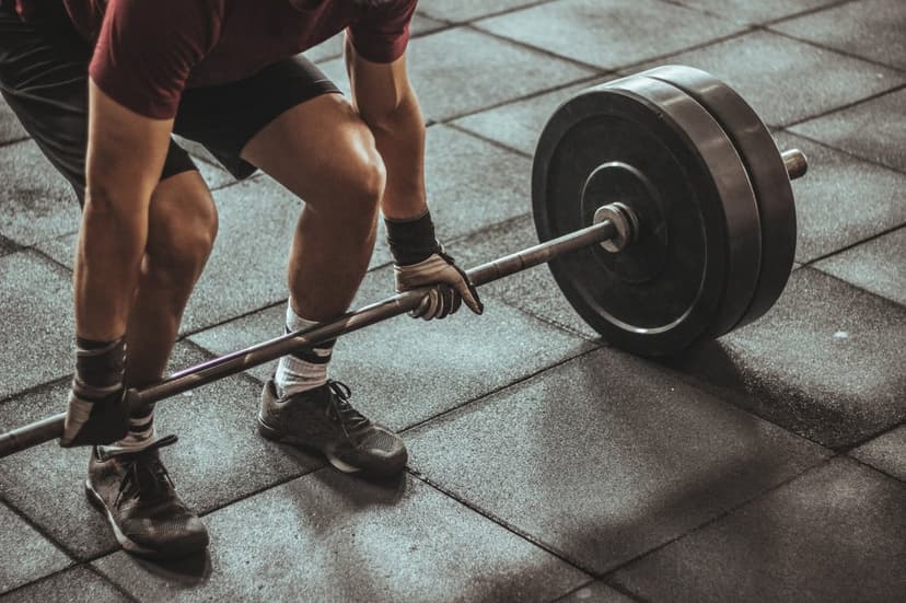 Muscular man wearing lifting gloves prepares to hoist a heavy barbell from a textured black rubber gym floor.