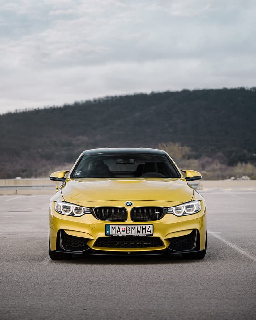 Yellow BMW M4 sits on asphalt with its kidney grille and sharp LED headlights prominent against a hilly background.