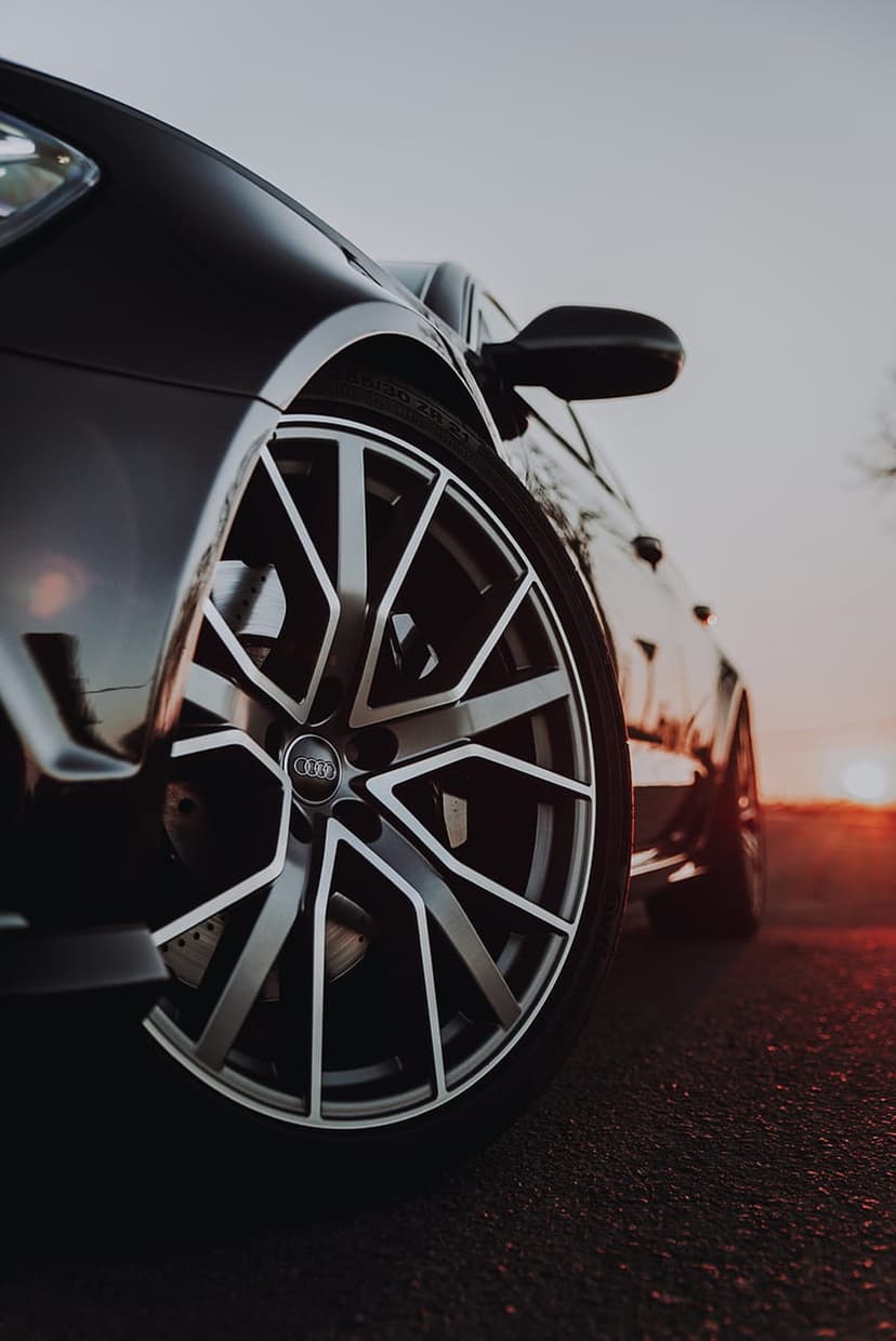 Audi alloy wheel with a black rim sits on asphalt during sunset, reflecting warm orange light and shadows.
