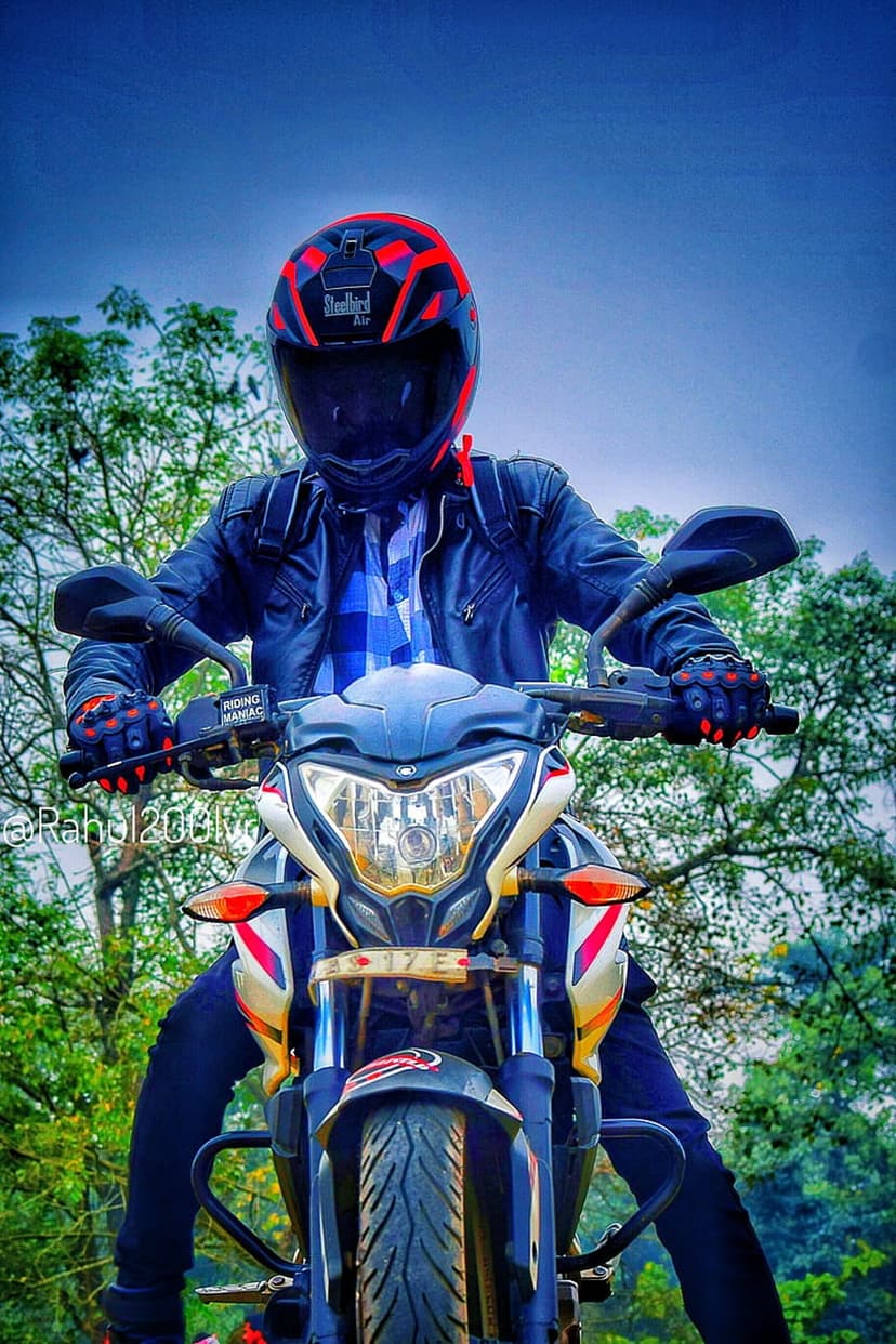 Motorcyclist in a red and black helmet and leather jacket sits on a bike against a blue sky and green trees.