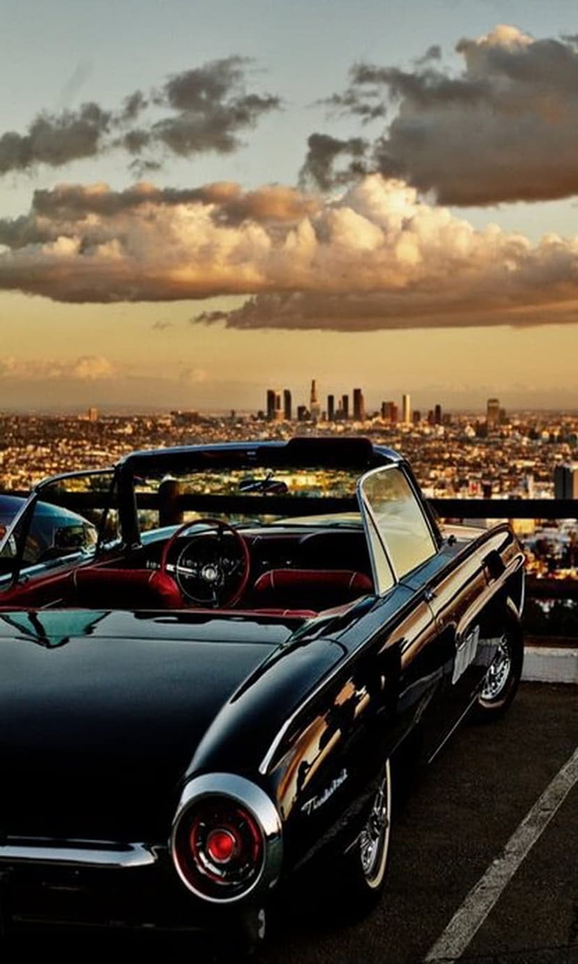 Black vintage convertible car parked on a hill overlooking the glowing Los Angeles skyline during a golden sunset.