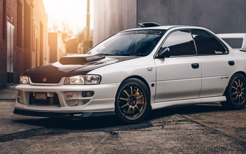 White Subaru Impreza WRX STI with a carbon fiber hood and gold wheels parked in a sunlit city alley.
