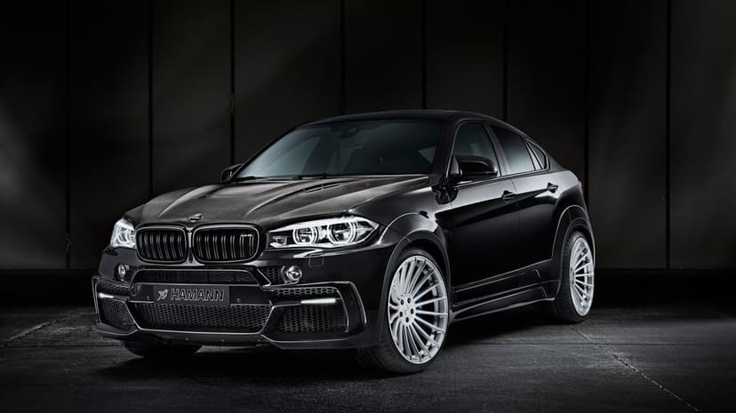 Black Hamann BMW X6 M with a carbon fiber hood and silver multi-spoke wheels positioned in a moody dark studio.