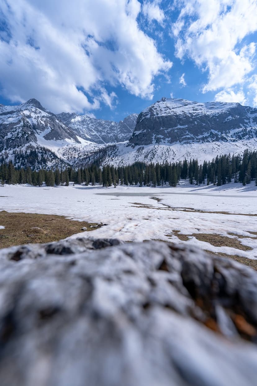 Snow-capped mountains stand behind a dense pine forest and partially frozen lake under a bright blue cloudy sky.