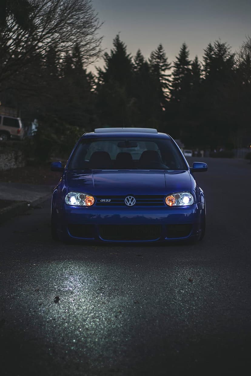 Blue Volkswagen R32 hatchback with glowing headlights on a wet asphalt road under a dark twilight sky.