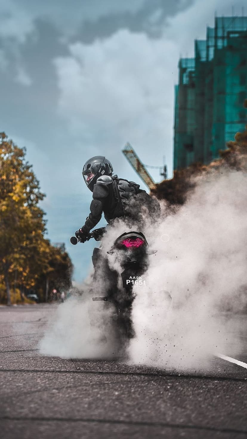 Motorcyclist performs a burnout with heavy white smoke and a pink glowing taillight in an urban street setting.