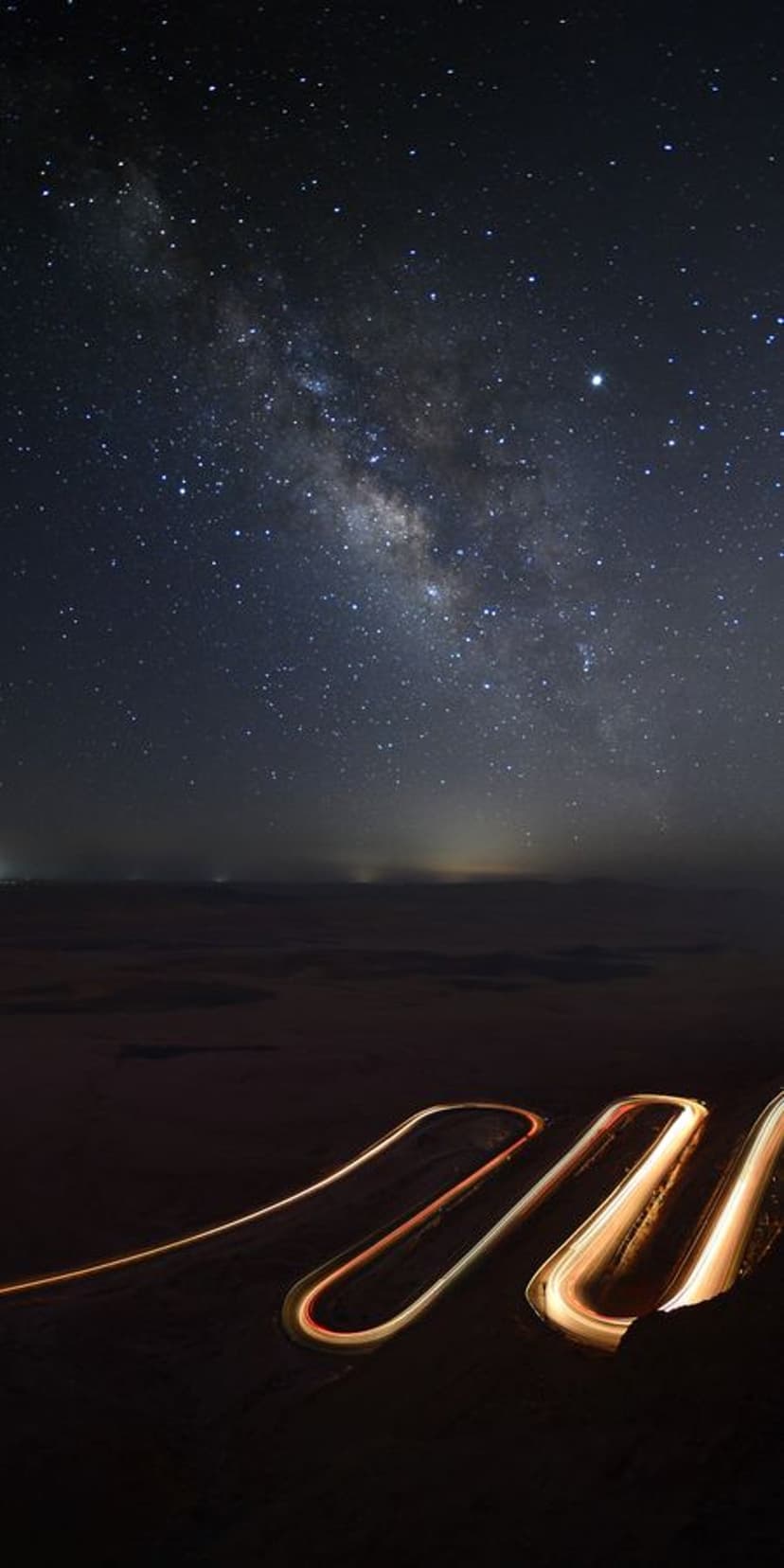 Winding road light trails glow beneath a star-filled Milky Way galaxy in a vertical night sky composition.