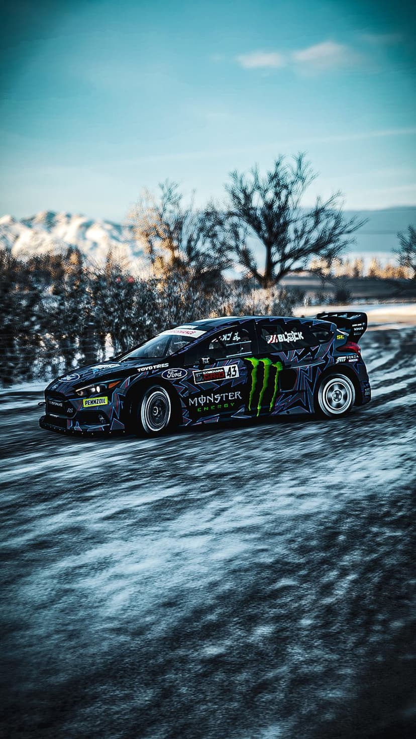 Monster Energy Ford Focus rally car drifting on a snowy track against a backdrop of blue skies and mountains.